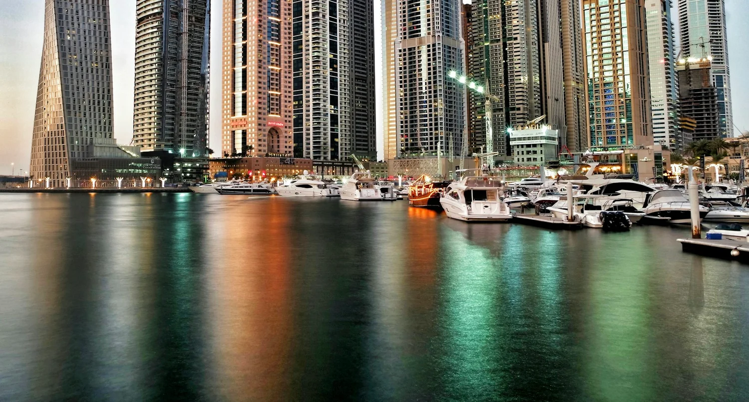 Real photograph of Dubai Marina waterfront and high-rise buildings reflected in the water.